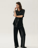 A high-waisted, straight-leg black silk pant. The pants have a smooth texture and a relaxed fit. The model is styled with matching button down blouse and black loafers.