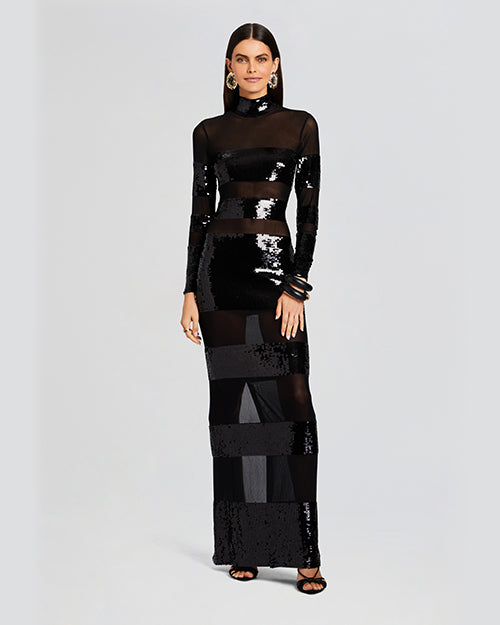 A full body view of a brunette model wearing a long-sleeved black dress with horizontal alternating mesh and sequin stripes. The dress features a high neckline and is paired with black heeled sandals, two thick black bangles, and round gold earrings. The background is plain white.