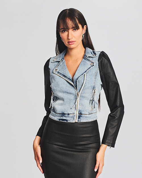 Front view of denim and leather jacket model with contrasting two-tone effect and the zipper closed. Jacket features silver hardware, styled with a form-fitting black dress.