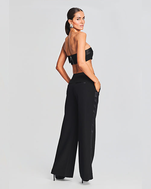 Back view of a model in a pair of black wide-leg pants with a high waist design and satin detail on waistband. The hem appears to be slightly flared. The model is styled with matching bandeau top.