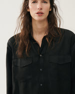 A close-up of a loose-fitting black button-up silk shirt with two chest pockets. The shirt has a collar, visible buttons down the front, and long sleeves.