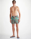 An image of a man standing wearing sunglass and swim shorts.  The turquoise men's swim shorts has dark red shadowed with neon orange drawings of an array of seaweed, kelp, and octopuses' legs.  The top of the swim shorts has an elastic waistband with a drawstring tie. 