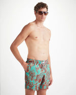 An image of a man standing from the knee up, wearing sunglasses, and swim shorts with his right hand in his side pocket. 
 The turquoise men's swim shorts has dark red shadowed with neon orange drawings of an array of seaweed, kelp, and octopuses' legs.  The top of the swim shorts has an elastic waistband with a drawstring tie. 