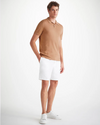 An image of a man standing wearing a camel color short sleeve polo shirt with a ribbed collar and two mother of pearl buttons, white knee length shorts, and white shoes. 