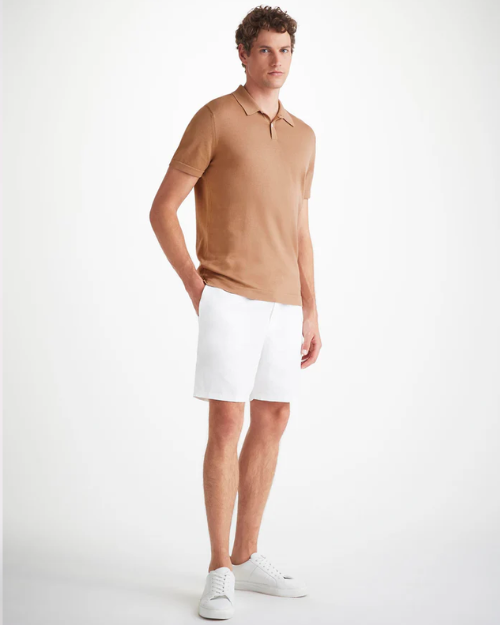 An image of a man standing wearing a camel color short sleeve polo shirt with a ribbed collar and two mother of pearl buttons, white knee length shorts, and white shoes. 