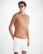 An image of a man from the knee up standing wearing a camel color short sleeve polo shirt with a ribbed collar and two mother of pearl buttons and white knee length shorts. The man has his right hand in his short pocket.