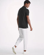 An image of a man standing backwards wearing a black color short sleeve polo shirt with a ribbed collar, white pants, and white shoes.