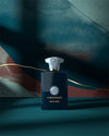 Dark blue fragrance bottle with light blue accents in front of elegant teal background. 