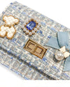 Up close image of the quilted ivory and gold bear charm, blue jewel with little pearls around charm, gold twist clasp, and a small blue bow with a pearl dangling off the middle of the bow on the front flap of the little girls blue tweed purse.