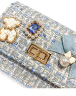 Up close image of the quilted ivory and gold bear charm, blue jewel with little pearls around charm, gold twist clasp, and a small blue bow with a pearl dangling off the middle of the bow on the front flap of the little girls blue tweed purse.