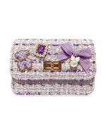 Purple tweed little girls purse with a quilted purple and gold bear charm, purple jewel with little pearls around charm, gold twist clasp, and a small purple bow with a pearl dangling off the middle of the bow on the front flap of the purse. 