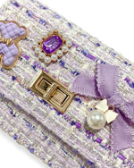 Up close image of the quilted purple and gold bear charm, purple jewel with little pearls around charm, gold twist clasp, and a small purple bow with a pearl dangling off the middle of the bow on the front flap of the little girls purple tweed purse. 