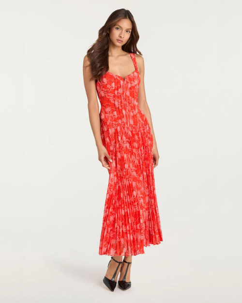 Person wearing a red floral pleated midi dress with thin straps and black heels on a plain background.