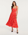 Person wearing a red floral pleated midi dress with thin straps and black heels on a plain background.