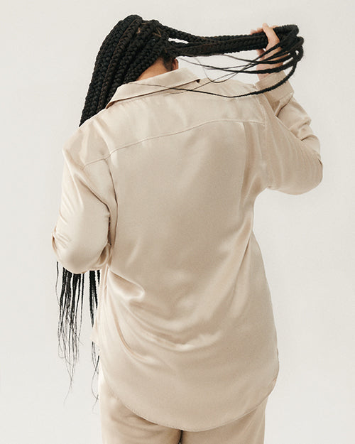 A knee-up backside view of a dark-haired model holding her hair up, wearing a beige button-down shirt featuring two chest pockets with flap closure, and matching bottoms. The background is plain grey.