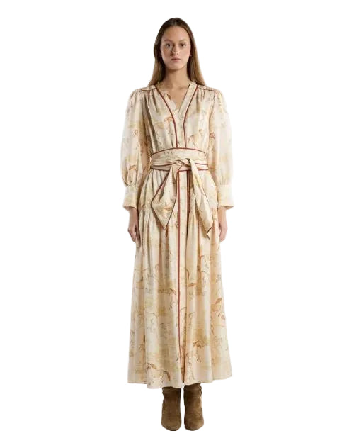 A woman wearing a long, beige dress with a subtle horse-themed print. The dress has long sleeves, a V-neckline, and a wide matching fabric belt tied at the waist, creating a gathered silhouette. 