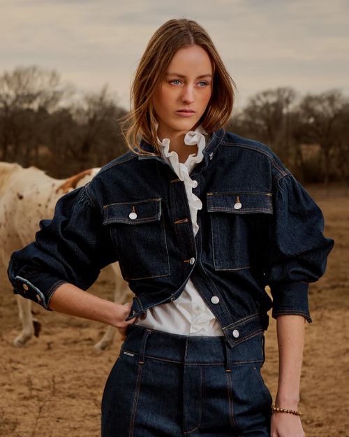 A woman in a denim jacket and pants stands beside a cow in a rural setting.