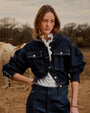A woman in a denim jacket and pants stands beside a cow in a rural setting.