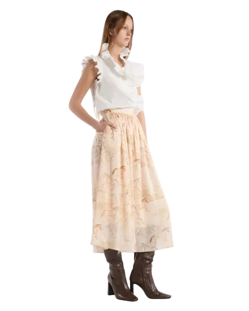 A woman wearing a white ruffle blouse with an elegant silk skirt with ruffles, set against a warm backdrop of painterly horses in soft desert tones.