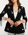 Person wearing a black embellished blazer with white lace-trim on a plain background.