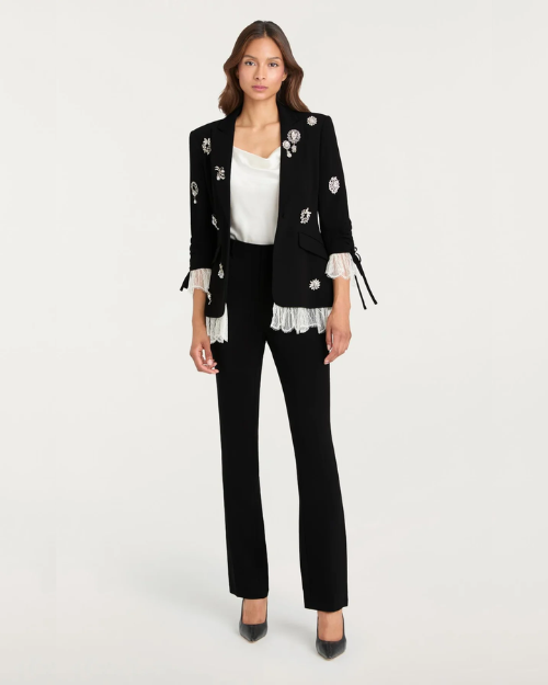 Person wearing a black embellished blazer with white lace-trim with black pants on a plain background.