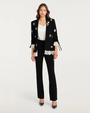 Person wearing a black embellished blazer with white lace-trim with black pants on a plain background.