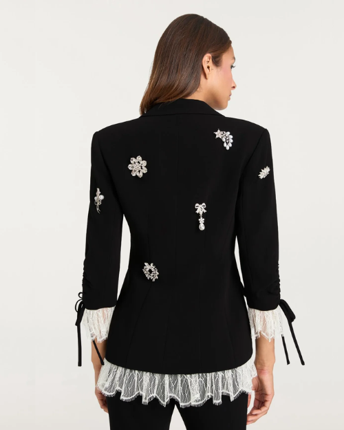 Back view of a person wearing a black embellished blazer with white lace-trim with black pants on a plain background.