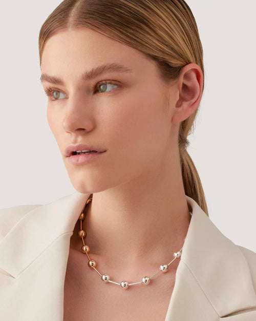 A model showcases a white blazer paired with a necklace that has luminous spheres that are paced out between elongated silver and gold beads. 