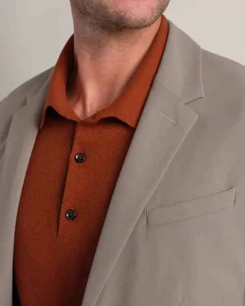 Close-up of a men's taupe tailored blazer layered over a rust knit polo shirt, highlighting the notch lapel and chest welt pocket.