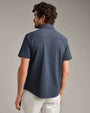 Back view of a navy short-sleeve button-down shirt with a classic collar, textured fabric, and tailored fit paired with light-colored pants.