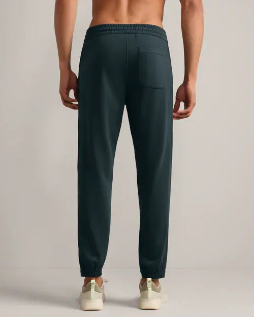 Back view of dark teal jogger pants featuring an elastic waistband, rear patch pocket and tapered cuffed legs, styled with white sneakers.