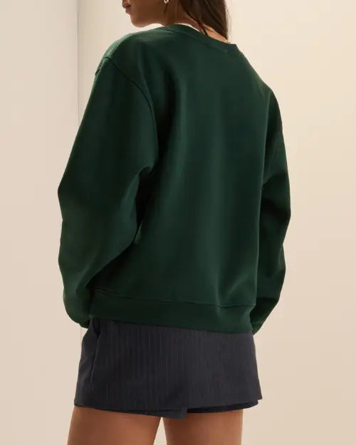 Back view of a dark green oversized crewneck sweatshirt with long sleeves and ribbed hem, styled with a navy pinstripe skort.