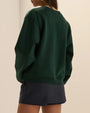 Back view of a dark green oversized crewneck sweatshirt with long sleeves and ribbed hem, styled with a navy pinstripe skort.