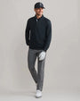 Person wearing a black quarter-zip performance pullover with long sleeves, gray golf pants, white golf shoes, and a navy cap in a studio setting.