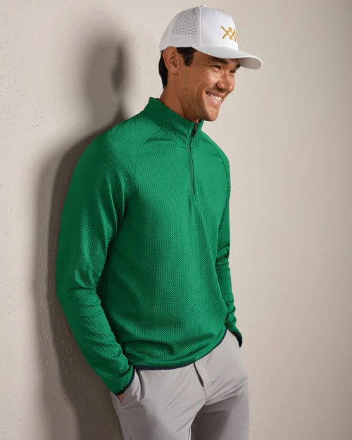 Person wearing a green textured quarter-zip performance pullover with long sleeves, light gray pants, and a white golf cap in a lifestyle setting.