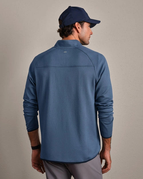 Back view of a blue textured quarter-zip performance pullover with long sleeves, athletic fit, and mock neck collar, paired with gray pants and a navy cap.