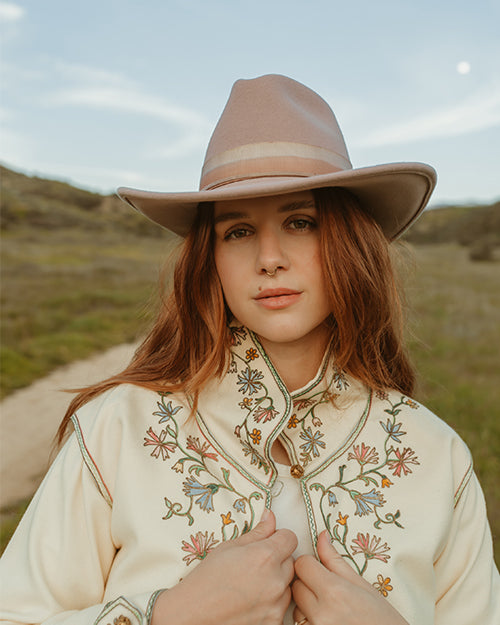 A model in an outside setting is wearing a wide-brimmed, mauve colored hat with a handcrafted detail around the base of the crown.