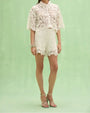 Women's white crochet lace blouse and matching lace shorts set with floral detailing, tie-neck collar, scalloped hems, and strappy heeled sandals.