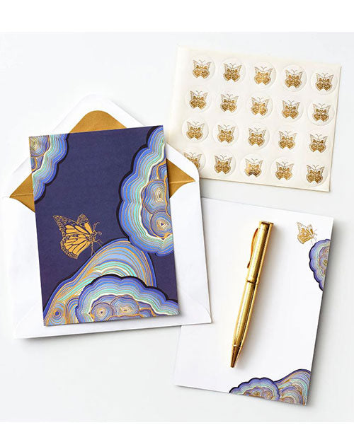 Stationery set with a blue notecard featuring a golden butterfly and abstract swirls, gold pen, sticker sheet with butterfly designs, and an envelope.