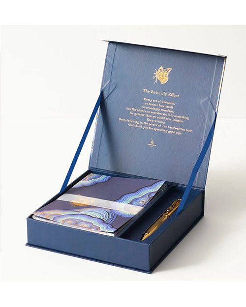 Open blue gift box with elegant stationery set, featuring a notebook with abstract patterns and a gold pen. Lid text reads "The Butterfly Effect." Elegant and inspiring.