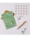 A floral notecard set includes a green card with blue and white flowers, matching envelope, flower stickers, a sheet for notes, and a gold pen.