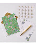 A floral notecard set includes a green card with blue and white flowers, matching envelope, flower stickers, a sheet for notes, and a gold pen.