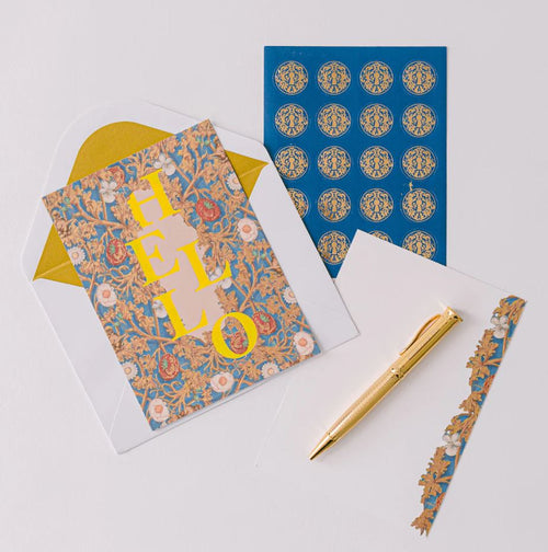 Elegant stationery set with a floral-patterned card that says "HELLO," gold pen, envelope, and decorative stickers. The tone is warm and inviting.