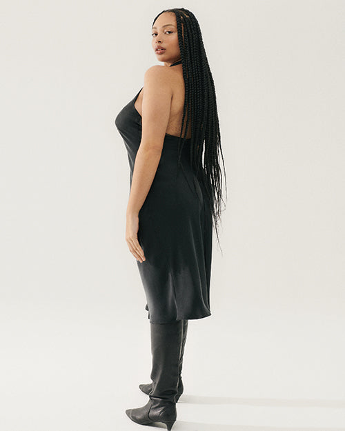 A back view of a sleeveless, halter neck midi dress in a black. The dress fits closely to the body, the back has a low cut and falls to just below the knee. Dress is paired with high black boots that rise to mid-calf.