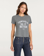 Person wearing a gray graphic T-shirt with “Cowgirls Do It Better” text and dark denim pants on a plain background.