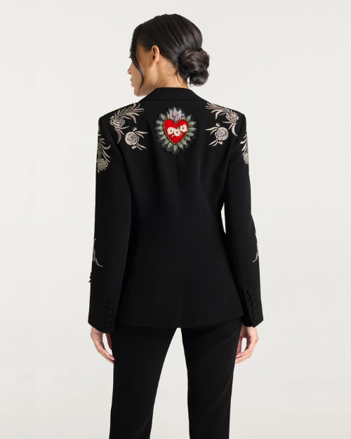 Woman wearing a black blazer with floral embellishments and a red heart patch against a white background. 