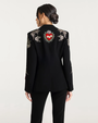 Woman wearing a black blazer with floral embellishments and a red heart patch against a white background. 