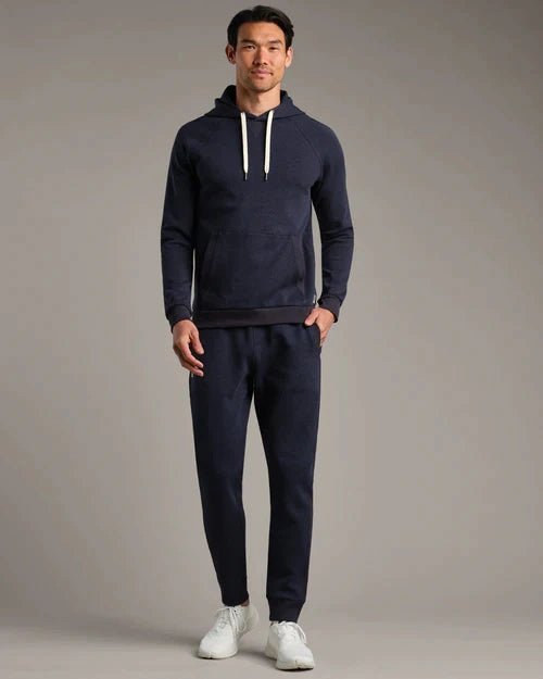 Person wearing a navy pullover hoodie and matching navy jogger pants with white drawstrings and white sneakers in a studio setting.