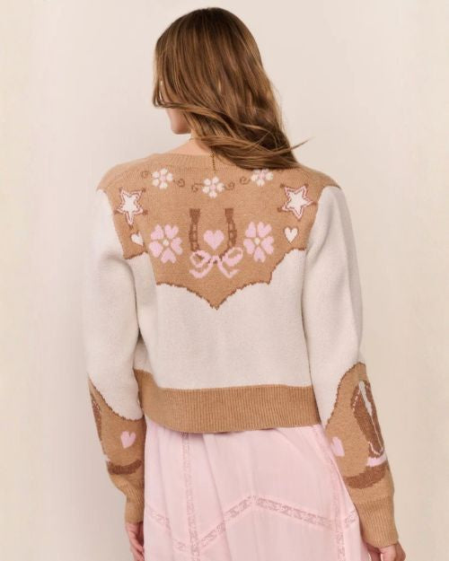 Back view of a person wearing a cropped tan and cream knit cardigan with star, floral, and horseshoe motifs, paired with a light pink dress.