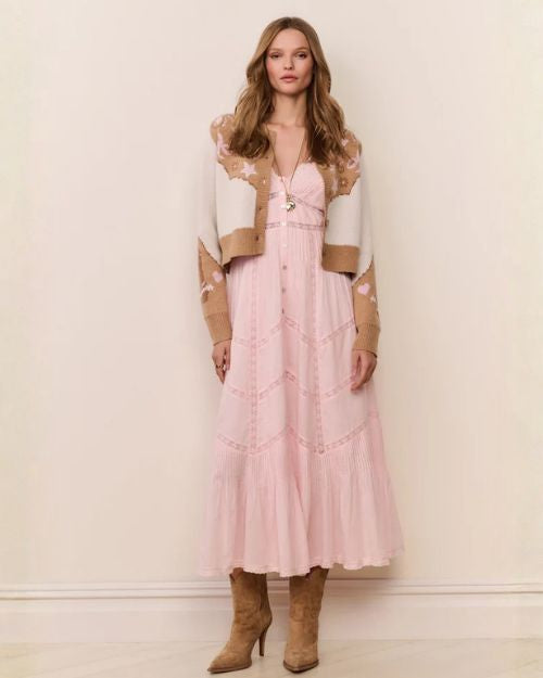 A woman wearing a long, light pink tiered dress with lace detailing, layered under a cropped tan and cream patterned cardigan, styled with a long pendant necklace and brown heeled boots.